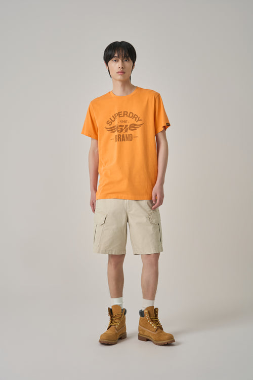 Person wearing an orange Superdry t-shirt with beige cargo shorts and brown boots on a plain background